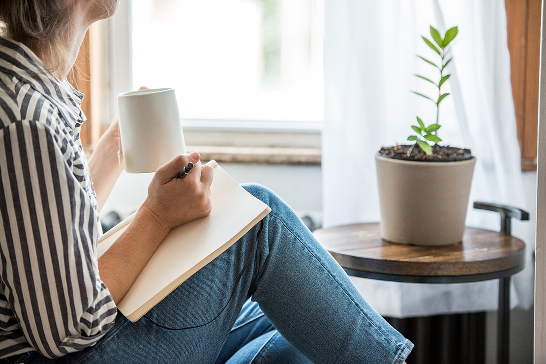 Frau sitzt alleine mit Tagebuch und Tasse Kaffee am Fenster – keine Angst, etwas zu verpassen