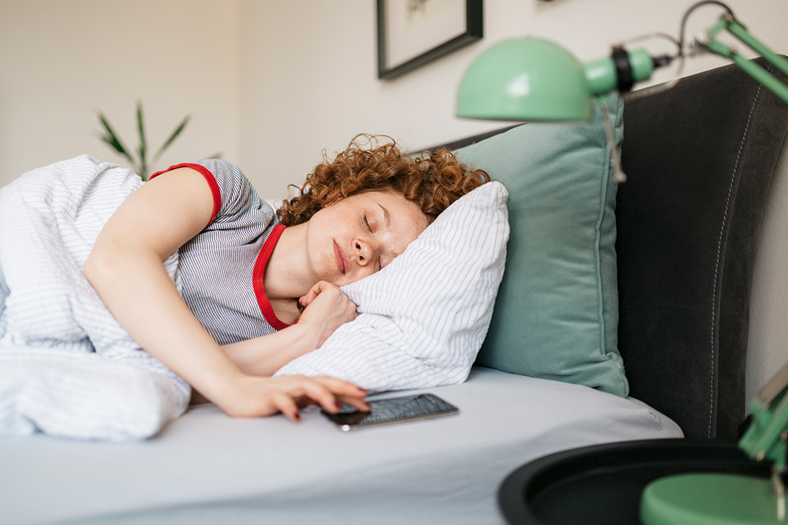 Frau liegt schlafend im Bett und drückt Handywecker auf Snooze 