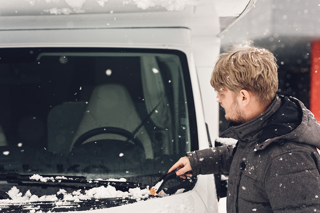 Mann kratzt Eis von Windschutz-scheibe von Wohnwagen in Vorbereitung zum Wintercamping